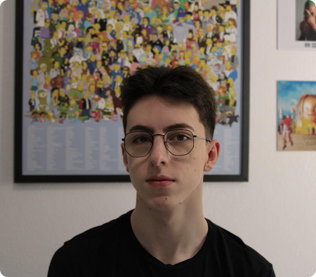 Retrato de um jovem de cabelos curtos e escuros, usando óculos de armação redonda e camiseta preta, posicionado em frente a uma parede branca com um quadro colorido ao fundo do desenho dos Simpsons